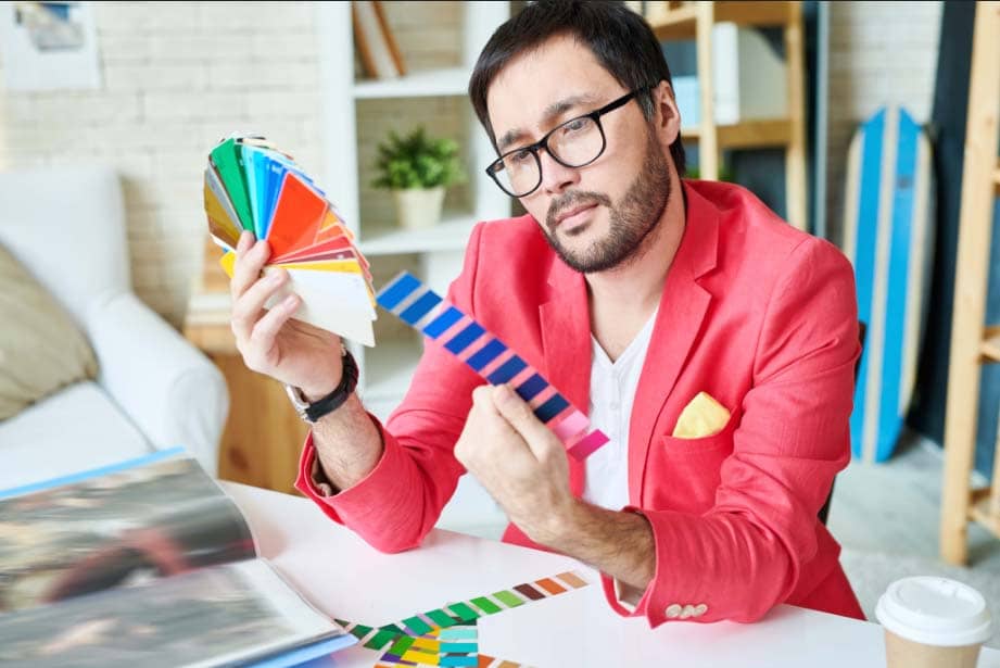 Confident young man working in atelier sitting at desk and choosing colors out of bright palettes.